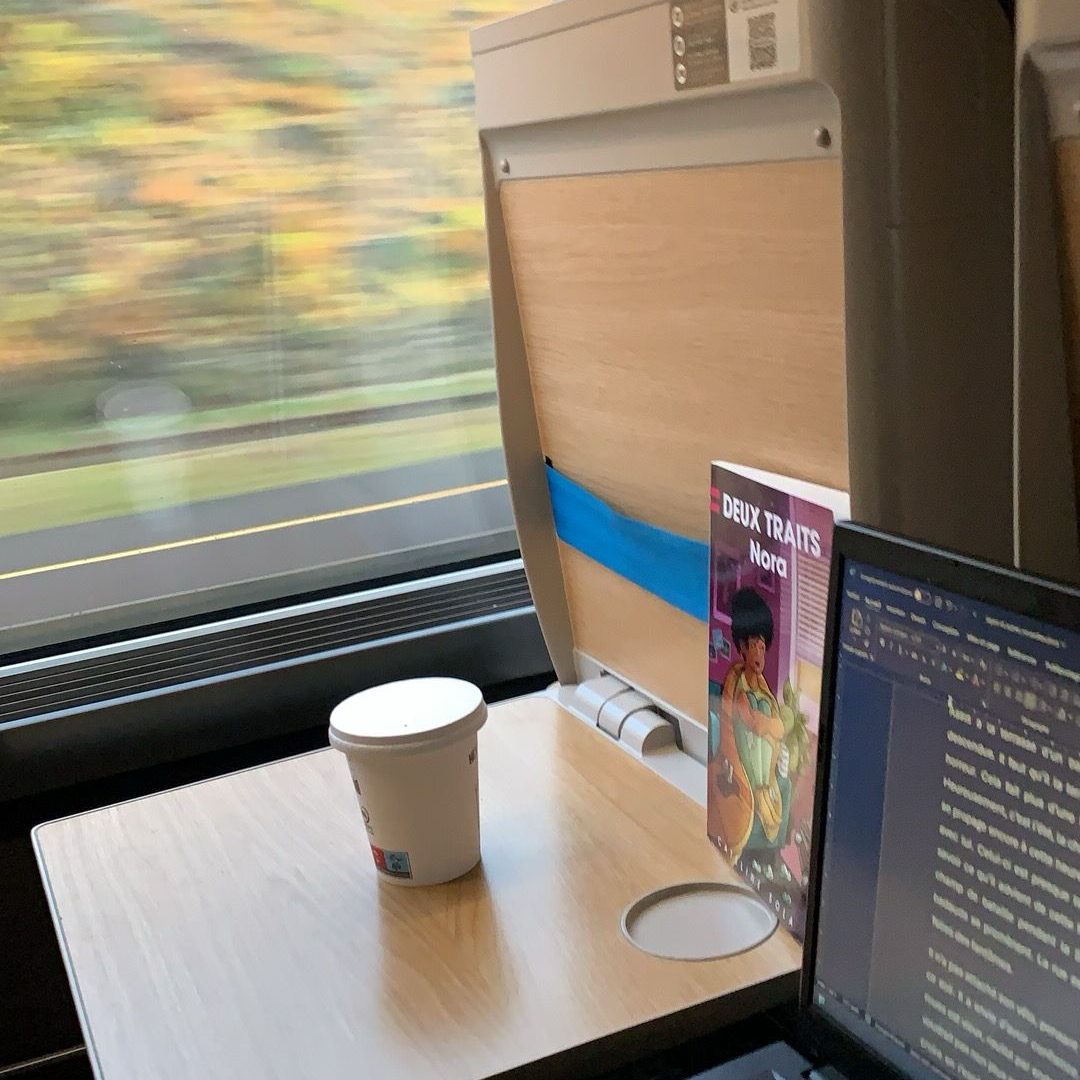 Table d'un train avec un ordinateur portable, une tasse et un livre à côté de la fenêtre.